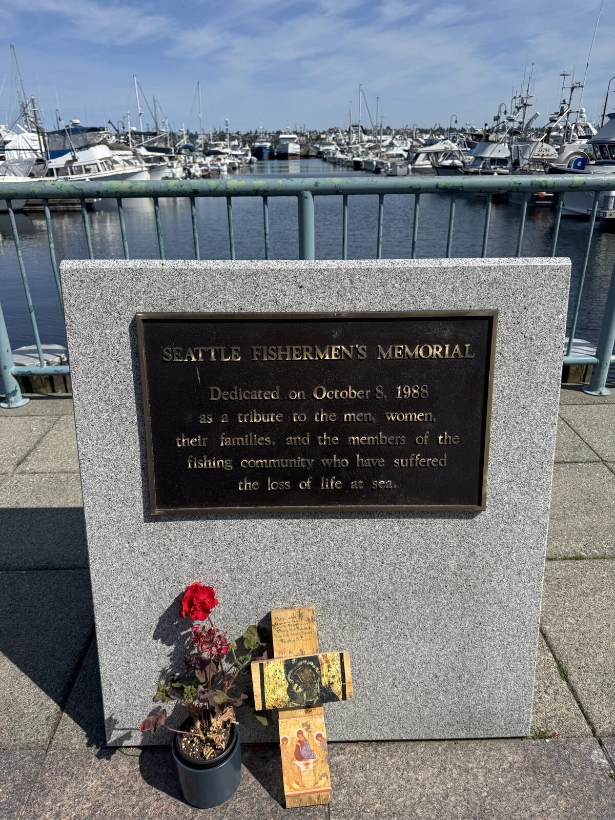 Seattle Fishermen's Memorial plaque honoring lives lost at sea, flowers at its base beside a marina full of boats